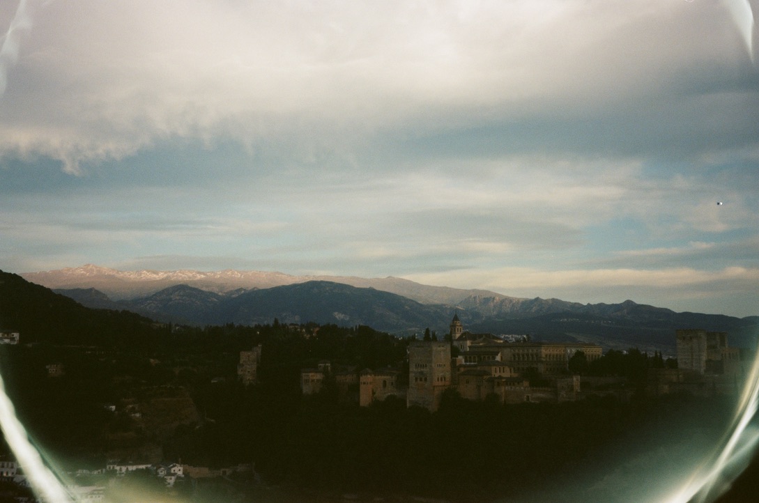 A palace and wooded hills beneath distant snowy mountains and a wide, clouded sky